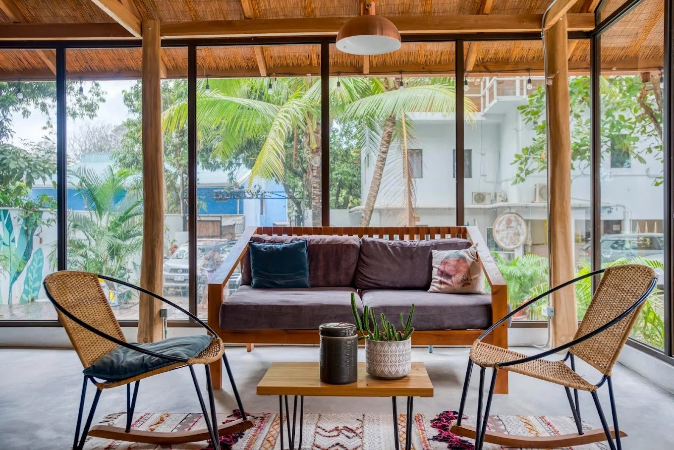 A relaxed lounge with a timber sofa, rattan chairs and palms framed through floor-to-ceiling glass